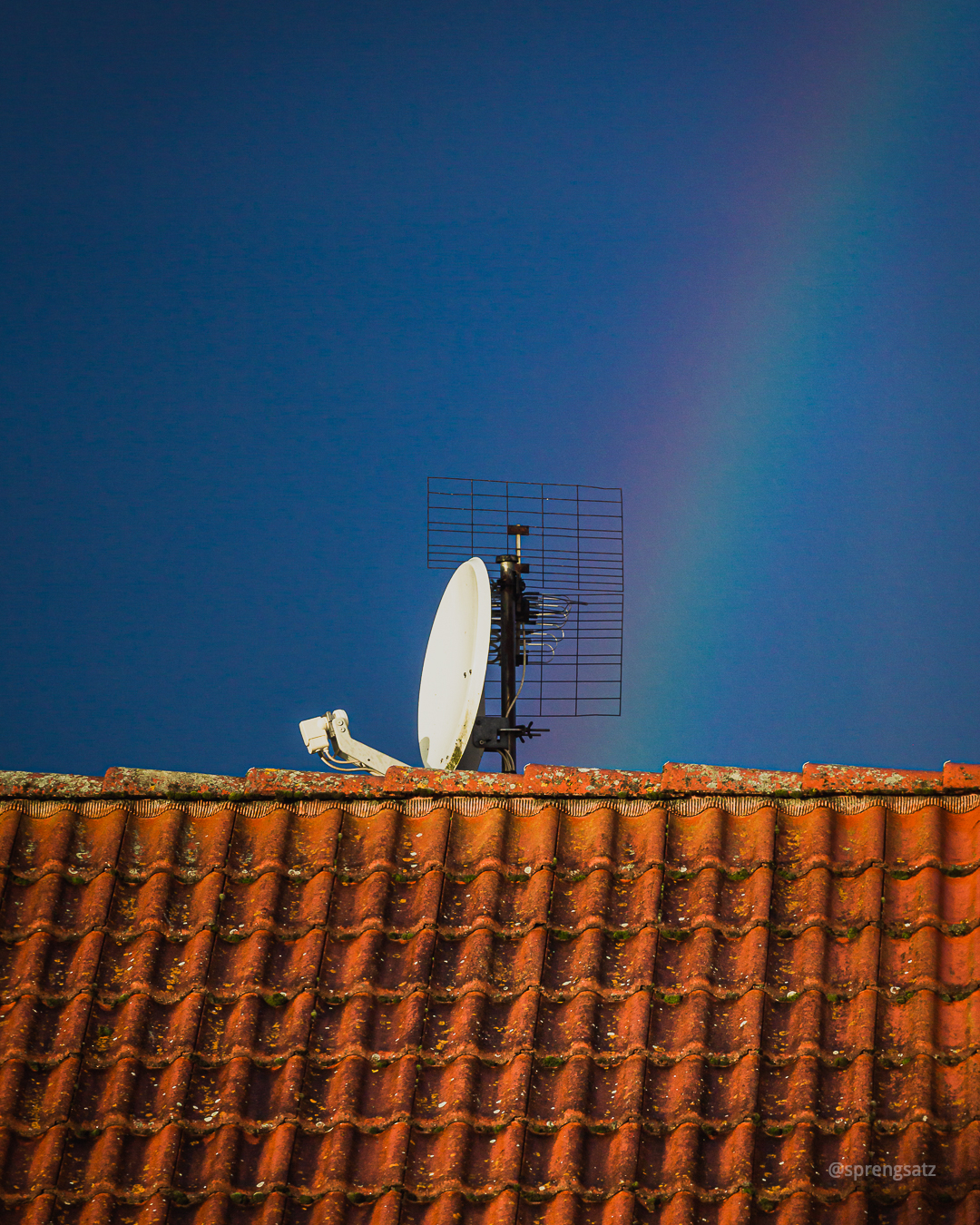 Hausdach mit SAT-Antenne vor blauem Himmel und Regenbogen